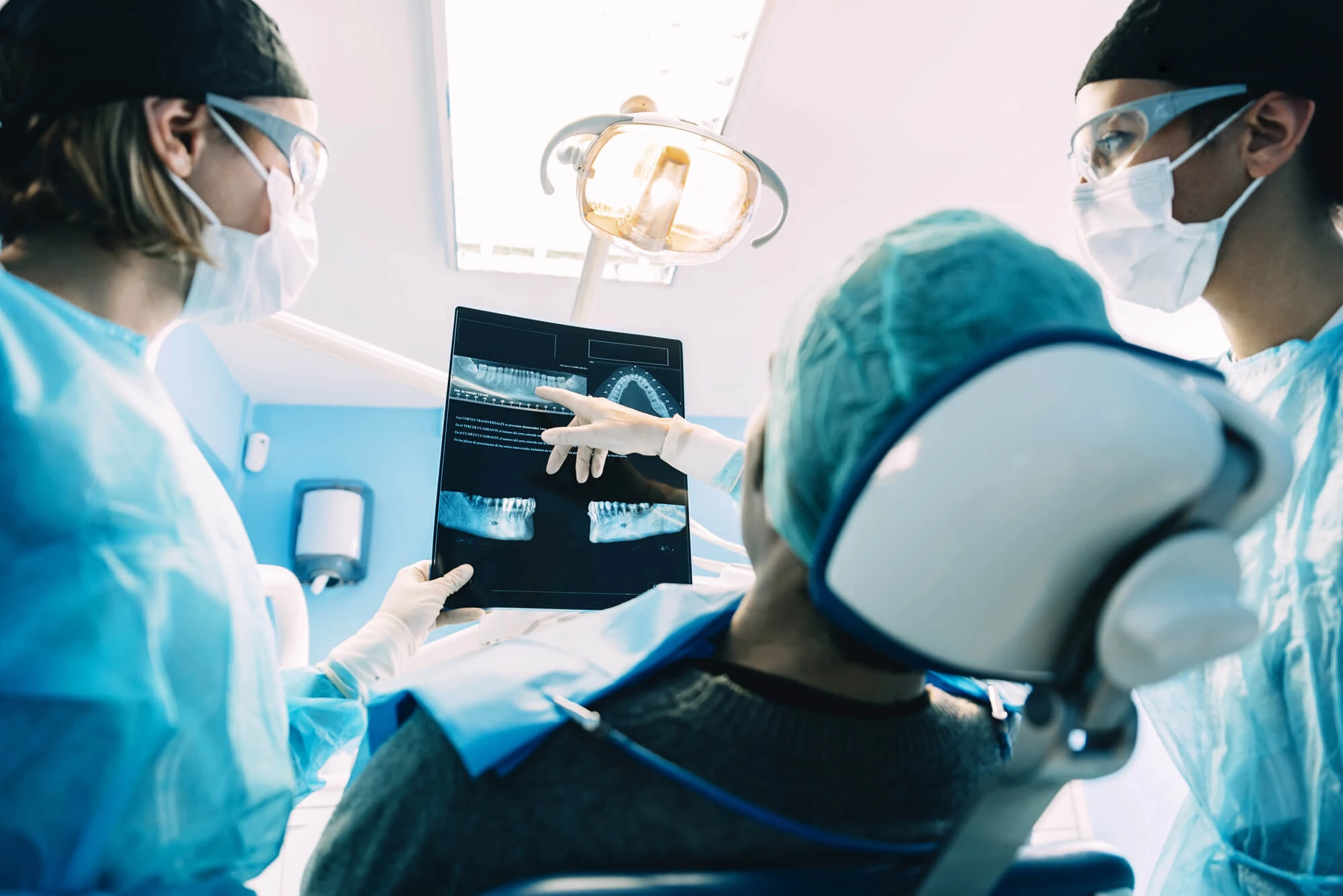 Two dentists wearing masks and gloves reviewing dental X-rays with a patient seated in a dental chair.