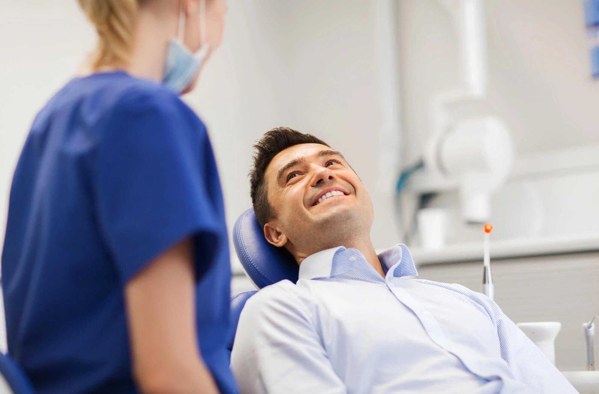 Smiling man reclined in a dental chair talking to a masked dental professional in blue scrubs.