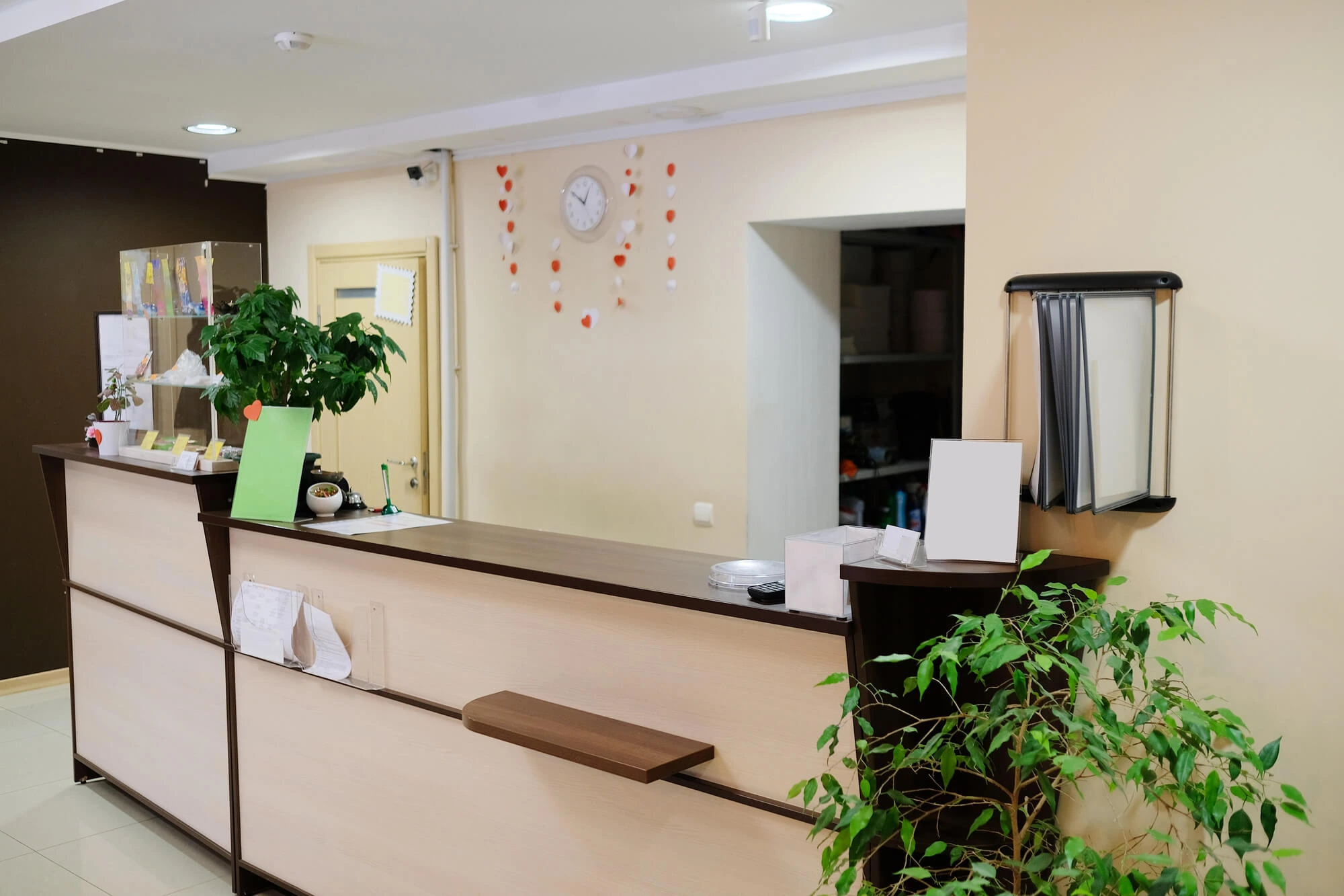 Reception desk area with potted plants, informational displays, and a clock on the beige wall decorated with red and white heart shapes.