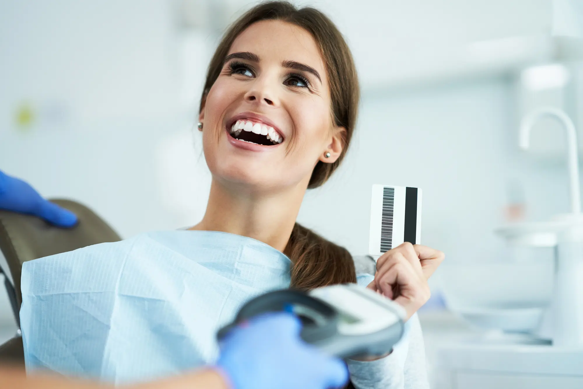 Smiling woman in dental chair holding a card while a gloved hand scans it with a payment terminal.
