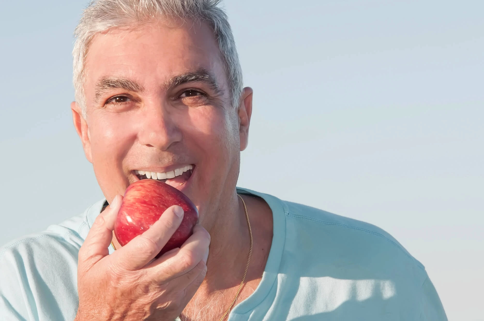 Middle-aged man with gray hair wearing a light blue shirt eating a red apple.
