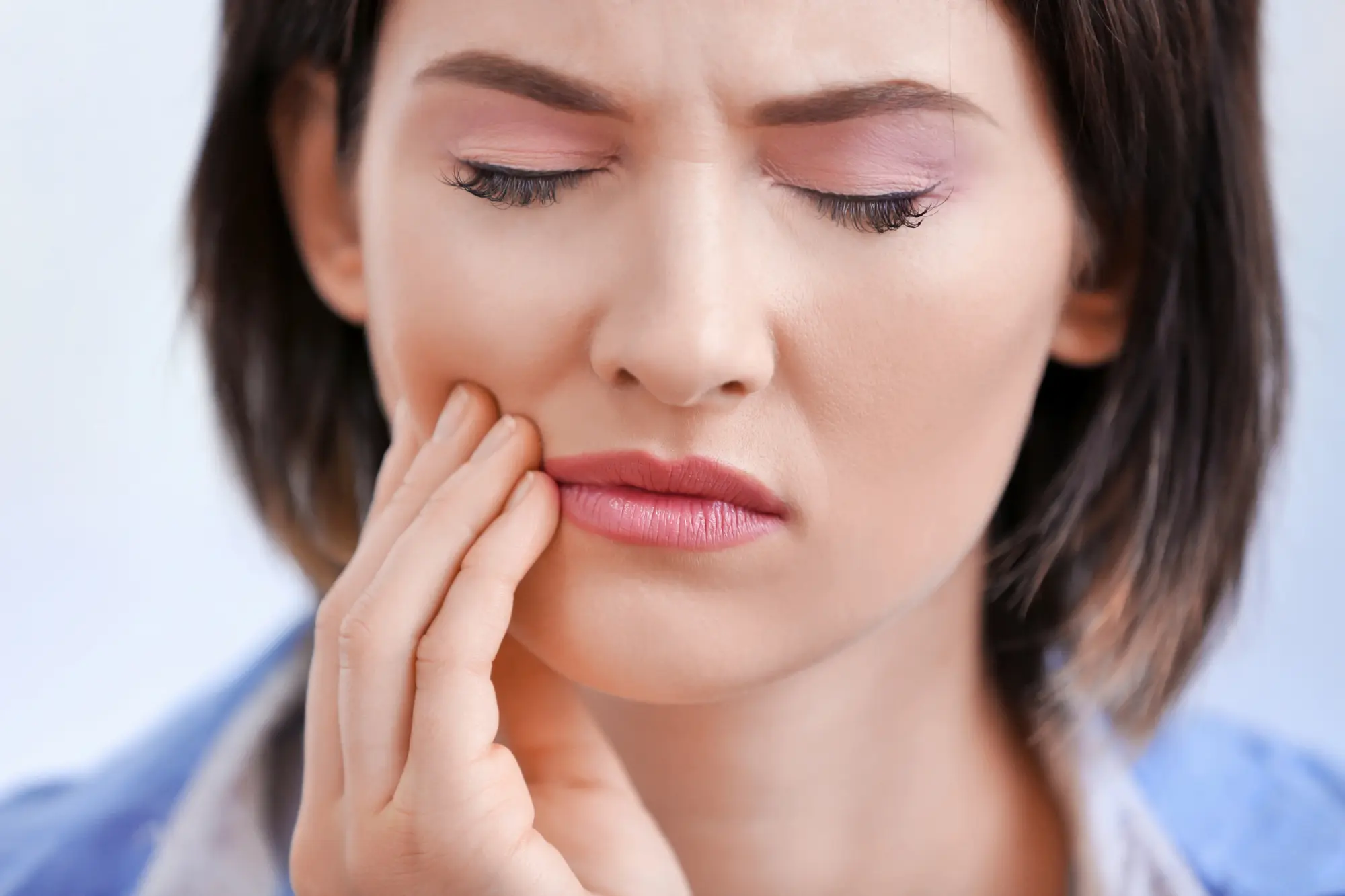 Close-up of a woman with closed eyes holding her cheek in pain, indicating a toothache or jaw discomfort.