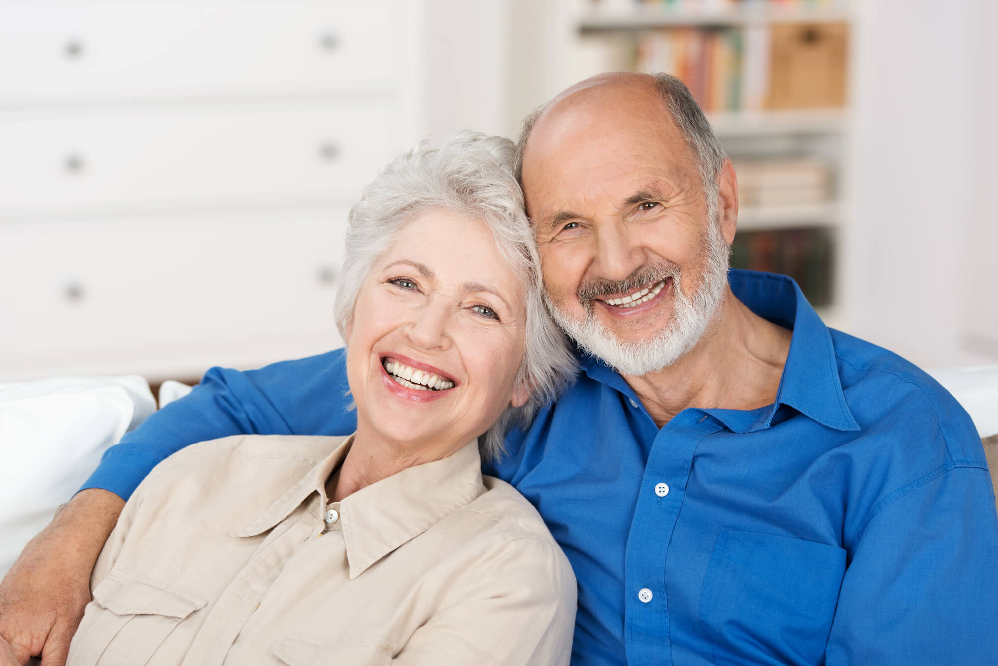 Smiling elderly couple sitting closely together on a couch in a bright living room.