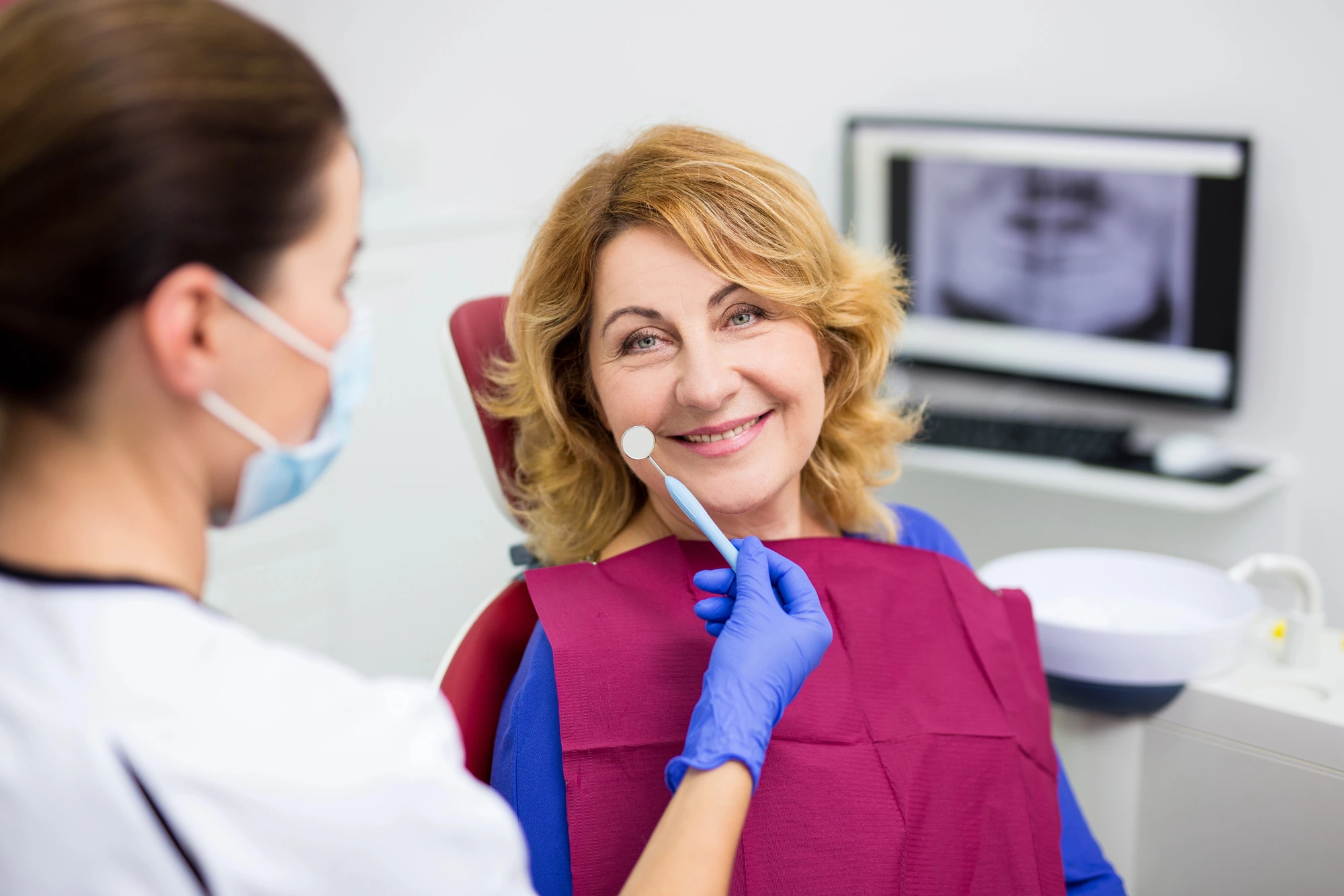Dentist wearing blue gloves and a mask holding a dental mirror near a smiling woman sitting in a dental chair.