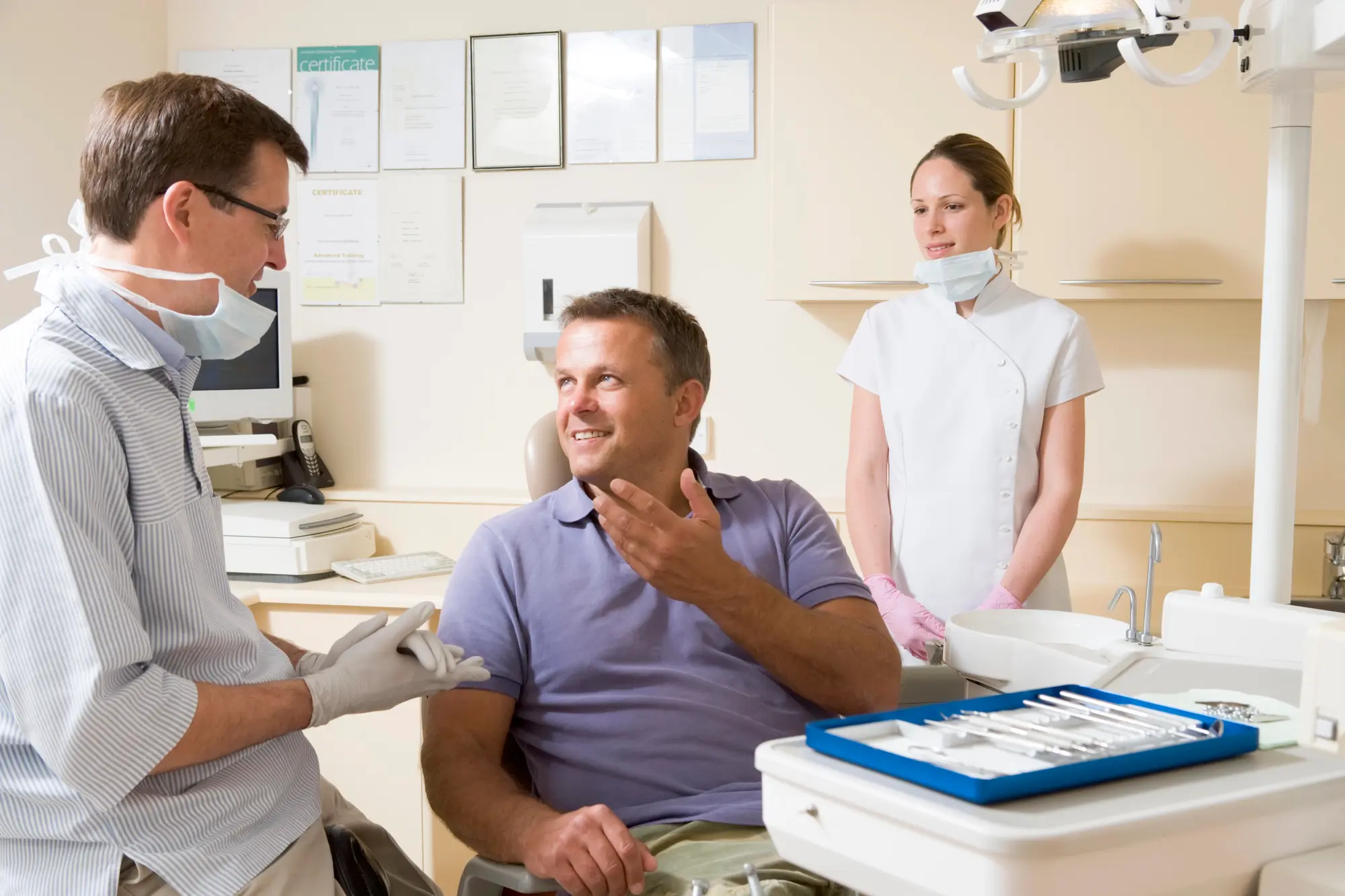 Dentist and dental assistant conversing with a smiling male patient seated in a dental chair with dental tools nearby.