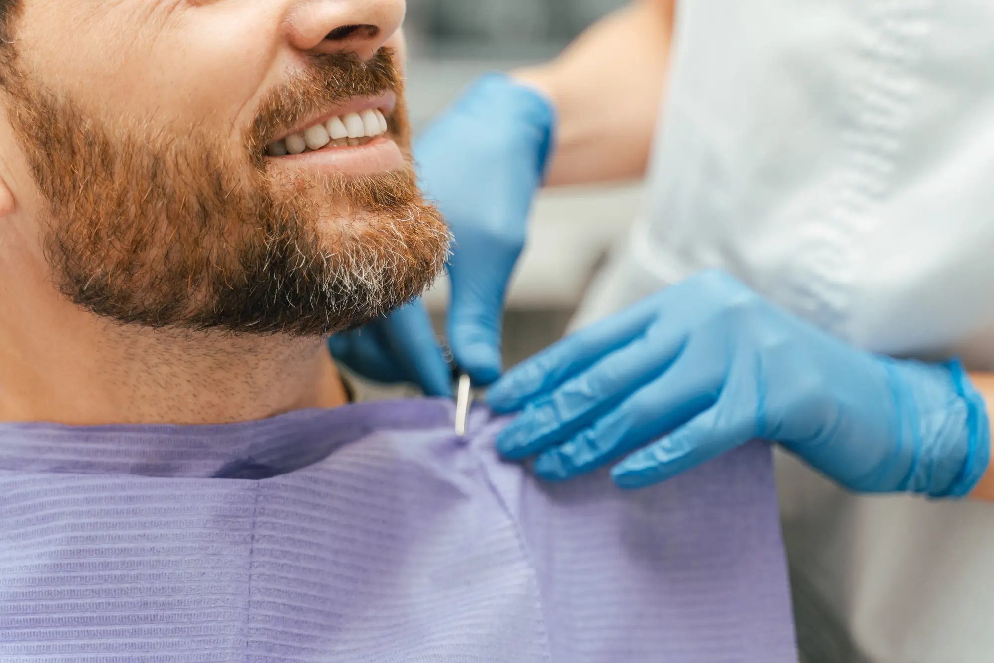 Smiling bearded man in a dental chair with a dental professional attaching a purple bib wearing blue gloves.