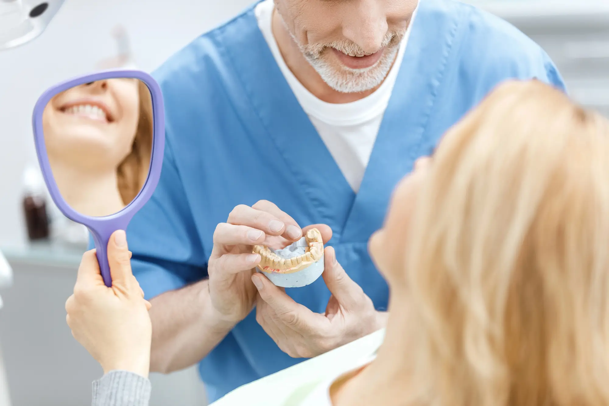 Dentist showing a dental mold to a smiling patient who is looking in a handheld mirror.