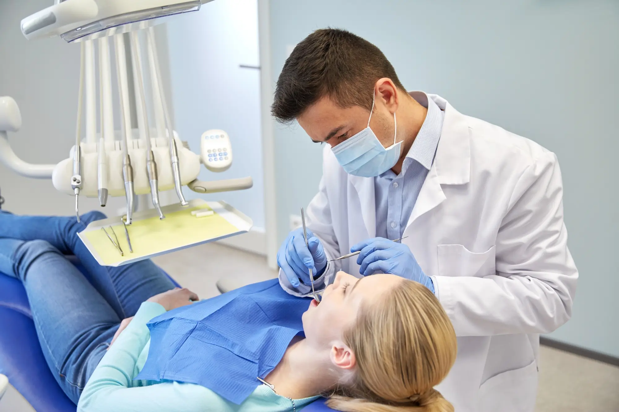 Dentist wearing mask and gloves examining a female patient's teeth in a dental clinic.
