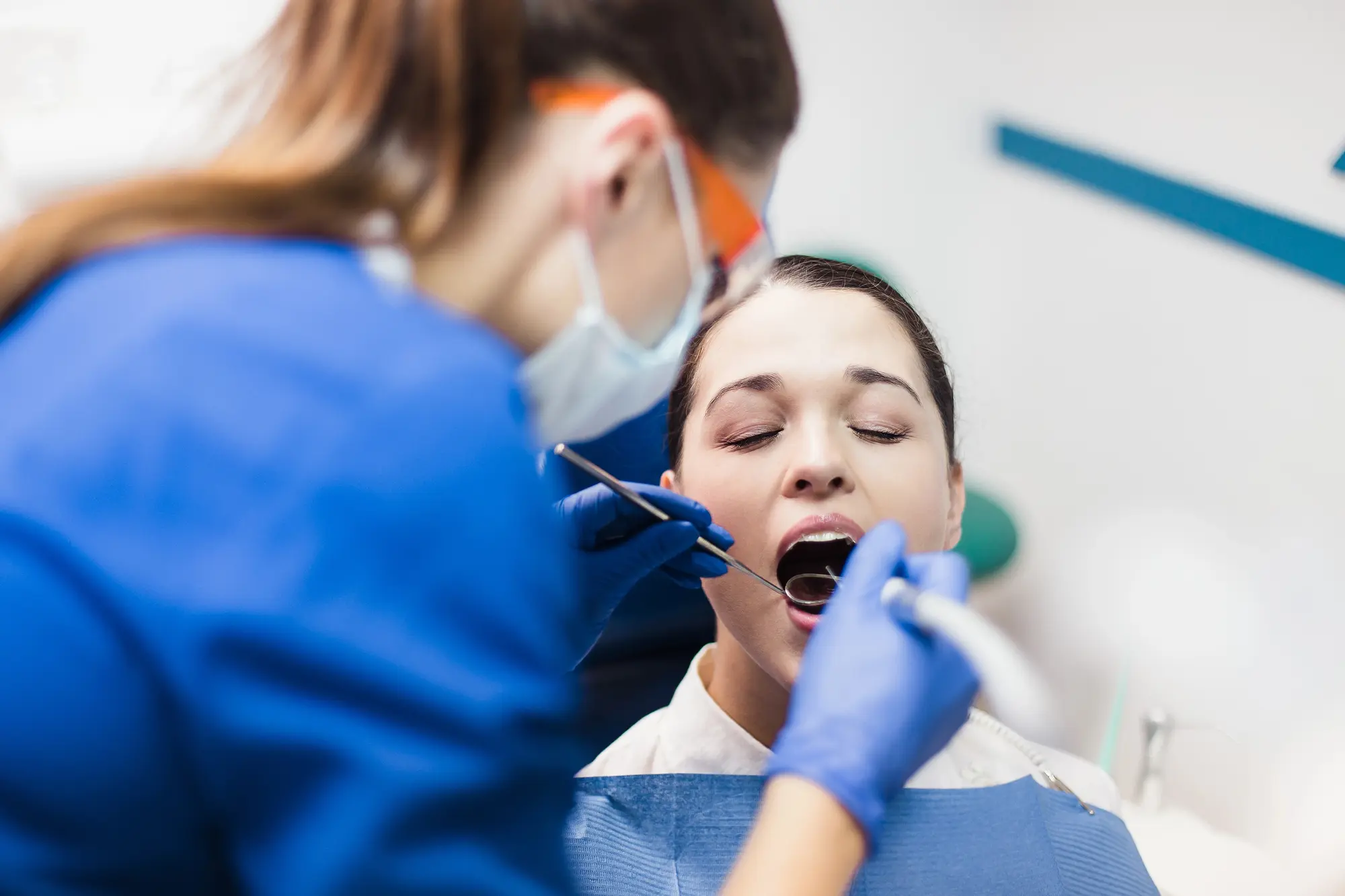 Dentist wearing gloves and a mask examining a female patient's open mouth with dental tools.