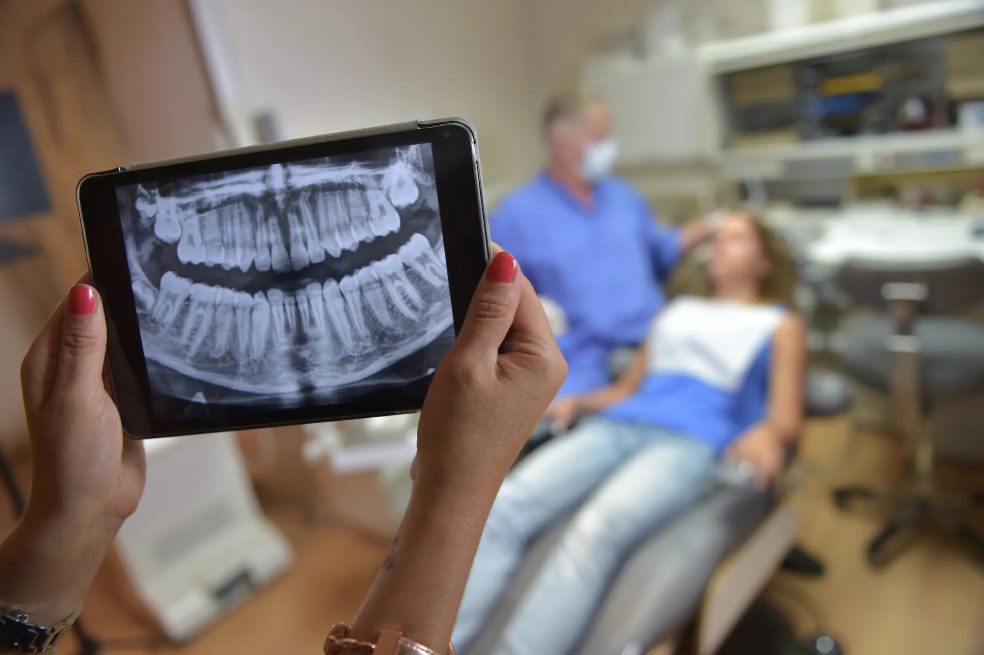 Hands holding a tablet showing a dental X-ray of teeth with a dentist and a patient in the background.