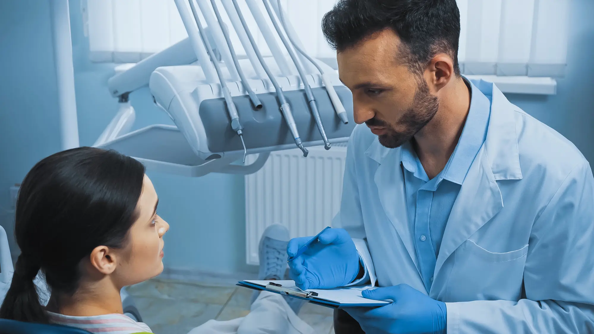 Dentist in a white coat and blue gloves discussing dental care with a seated female patient in a dental office.