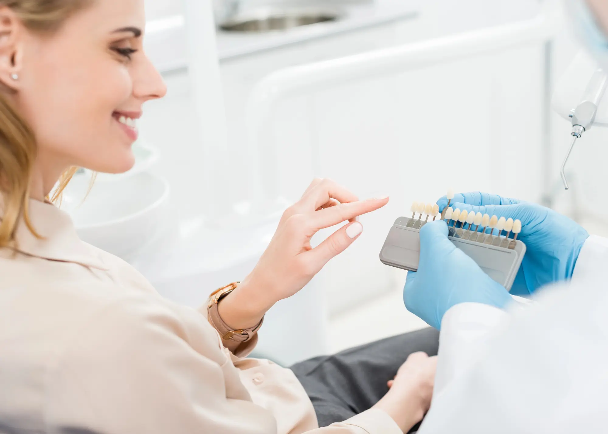 Woman smiling in dental clinic selecting a tooth shade from a shade guide held by a dentist wearing blue gloves.