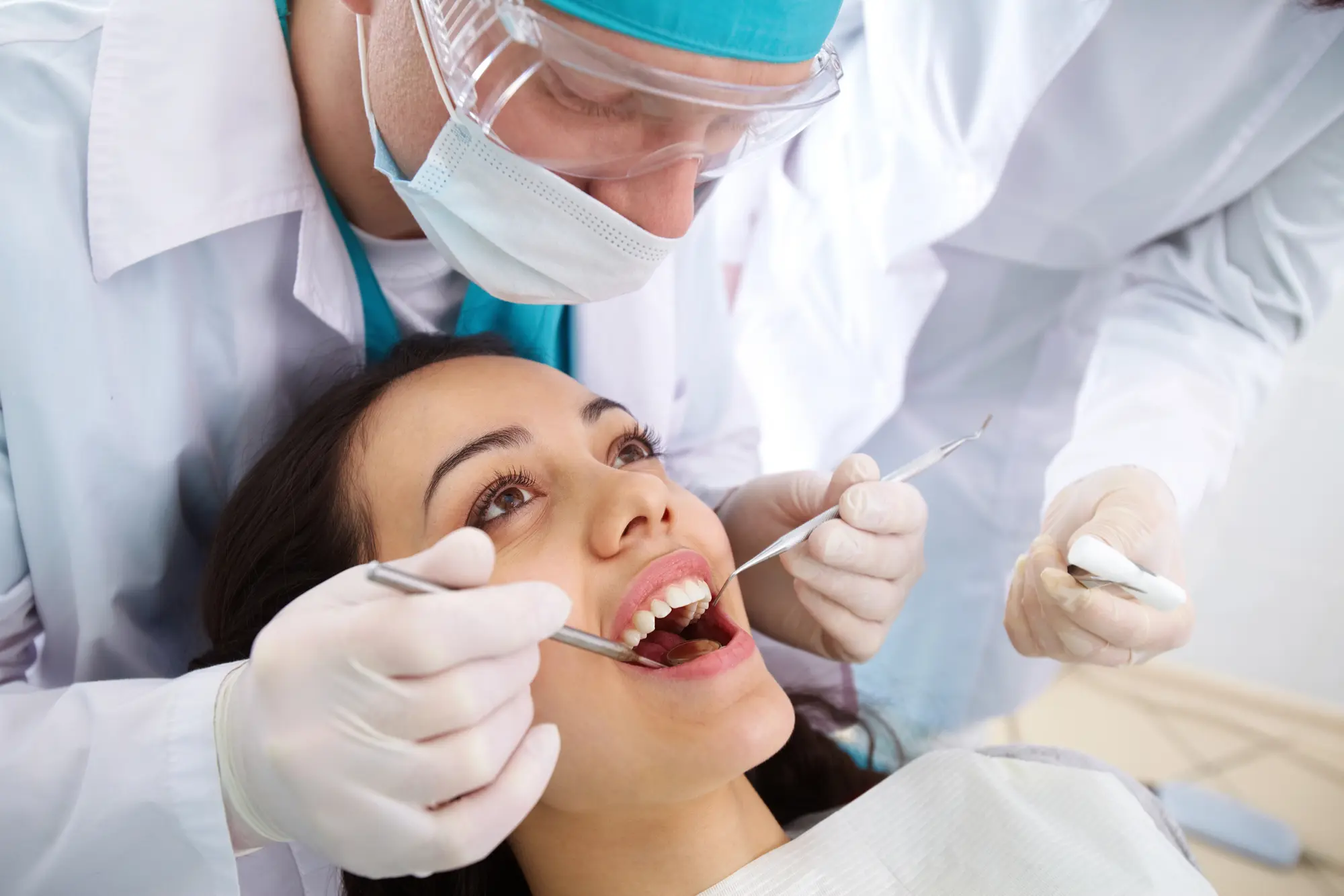 Dentist wearing protective glasses and mask examining a smiling woman's teeth with dental tools.