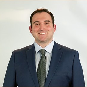 Smiling man in a dark suit, white shirt, and green tie against a plain light background.