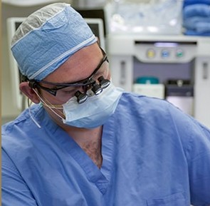 Surgeon wearing blue scrubs, surgical cap, mask, and magnifying glasses focused on a procedure.