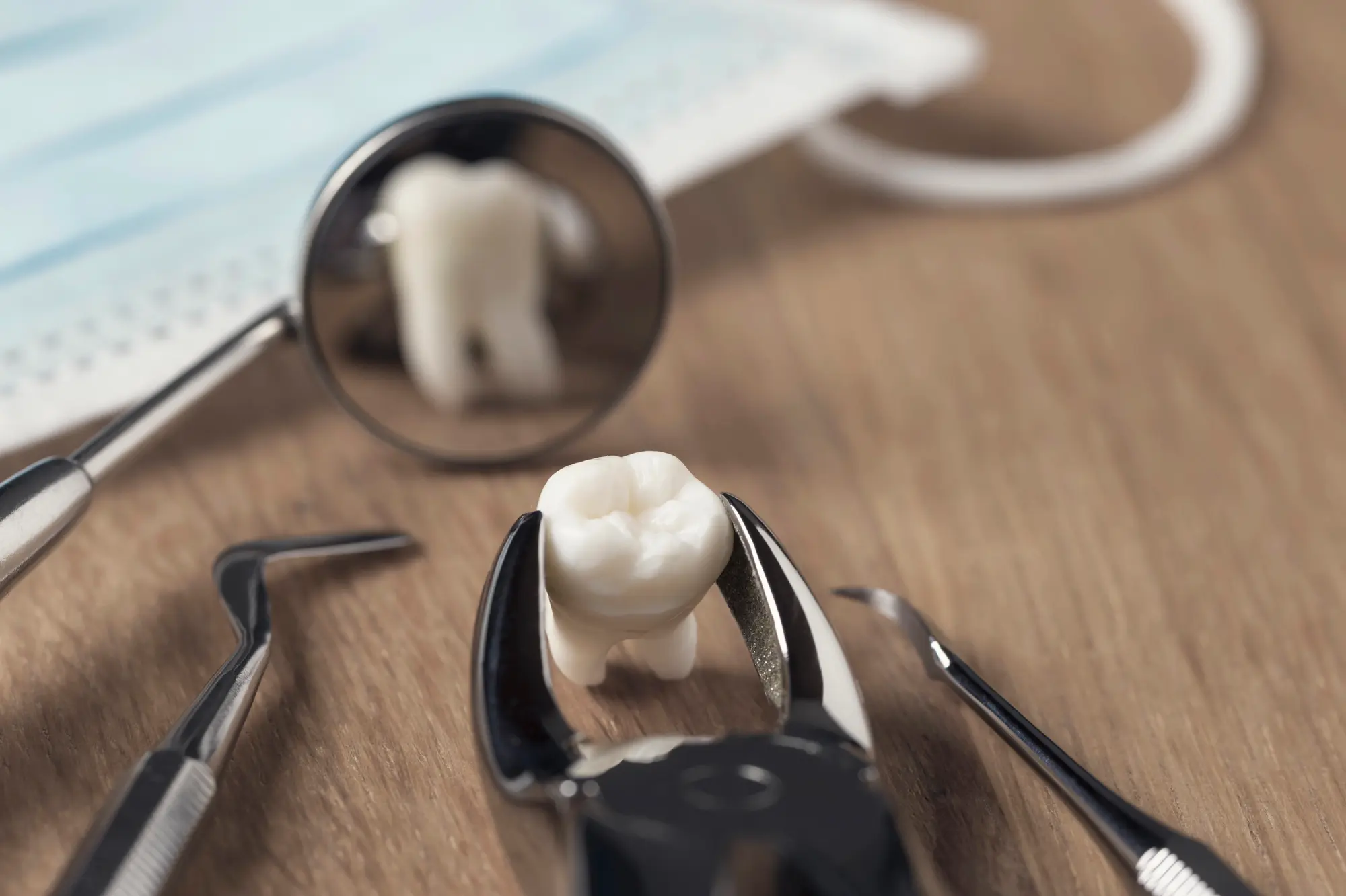 Dental tools including mirror, scaler, and forceps holding a model tooth, with a surgical mask in the background.
