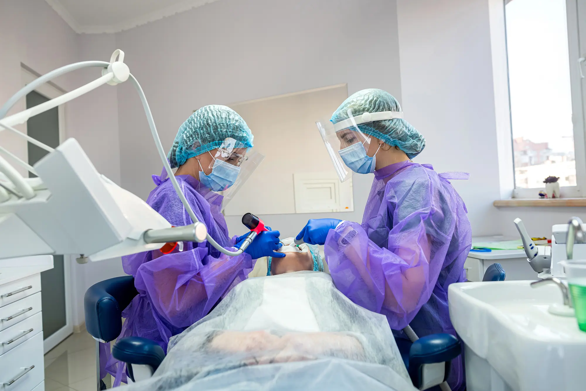 Two dentists wearing protective gear treating a patient lying in a dental chair in a clinic.