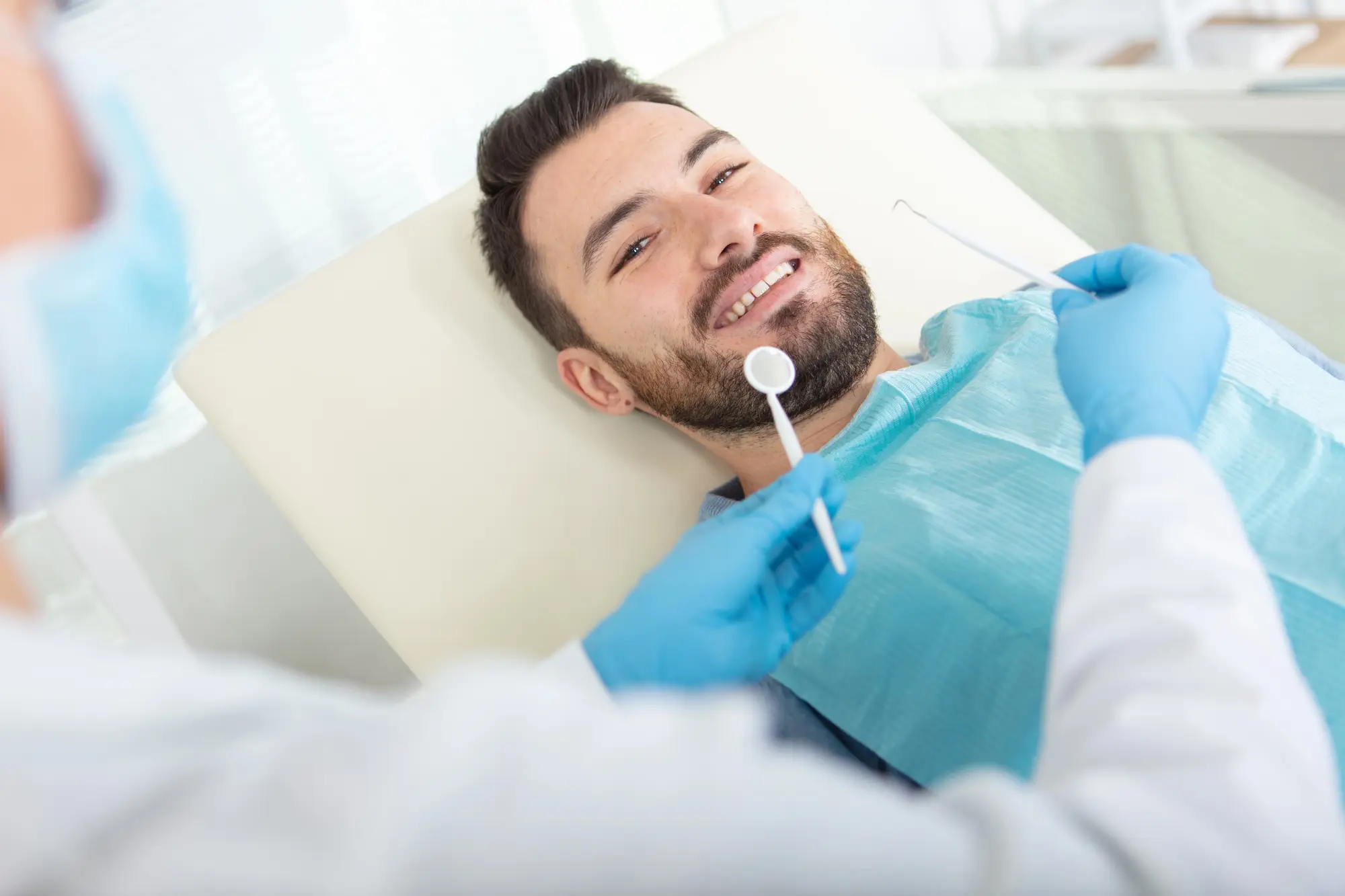 Smiling man lying on a dental chair wearing a blue bib while a dentist in blue gloves holds dental tools near him.