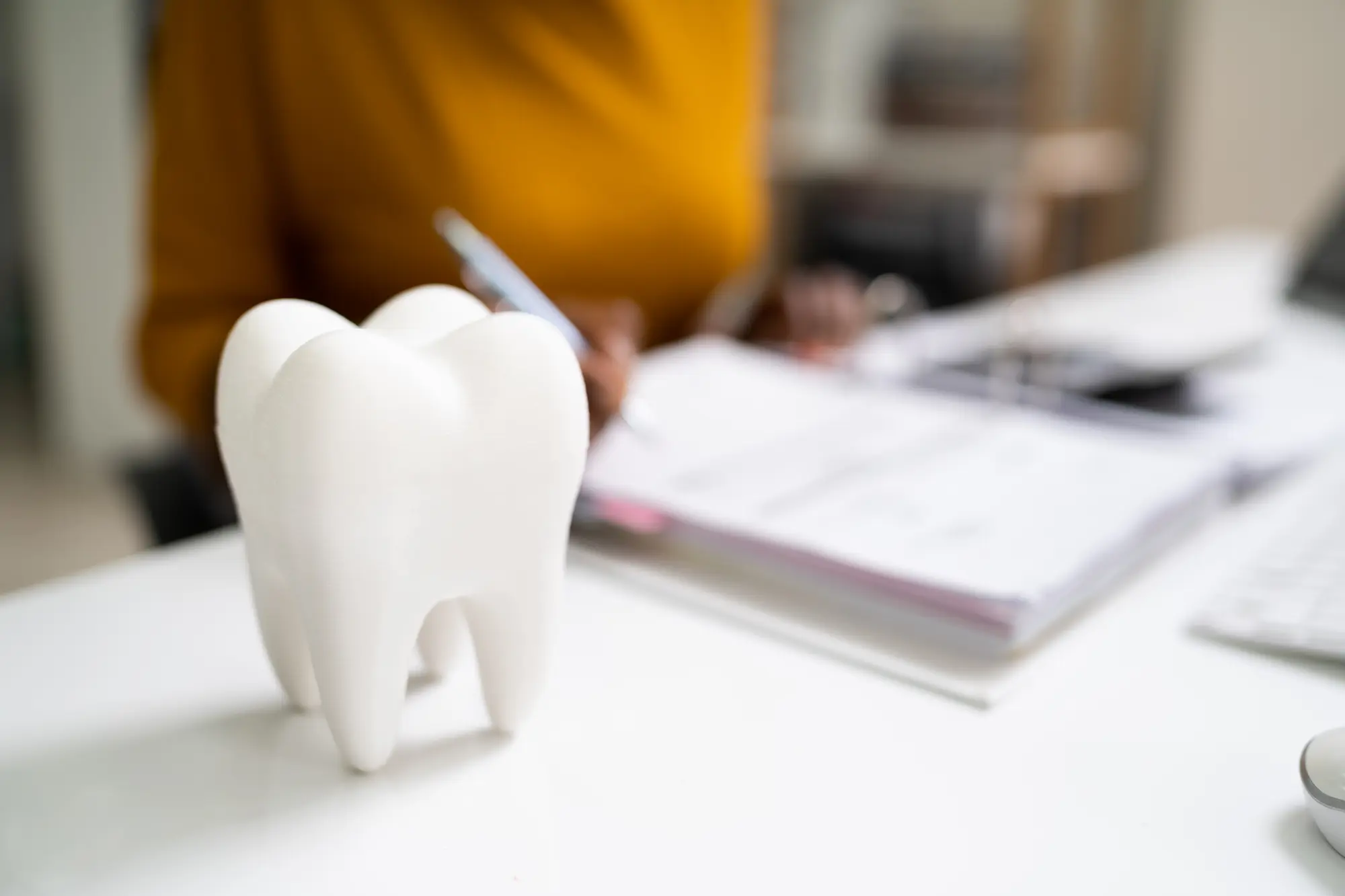 Large white tooth model on a desk with blurred person writing in background, symbolizing dental care.