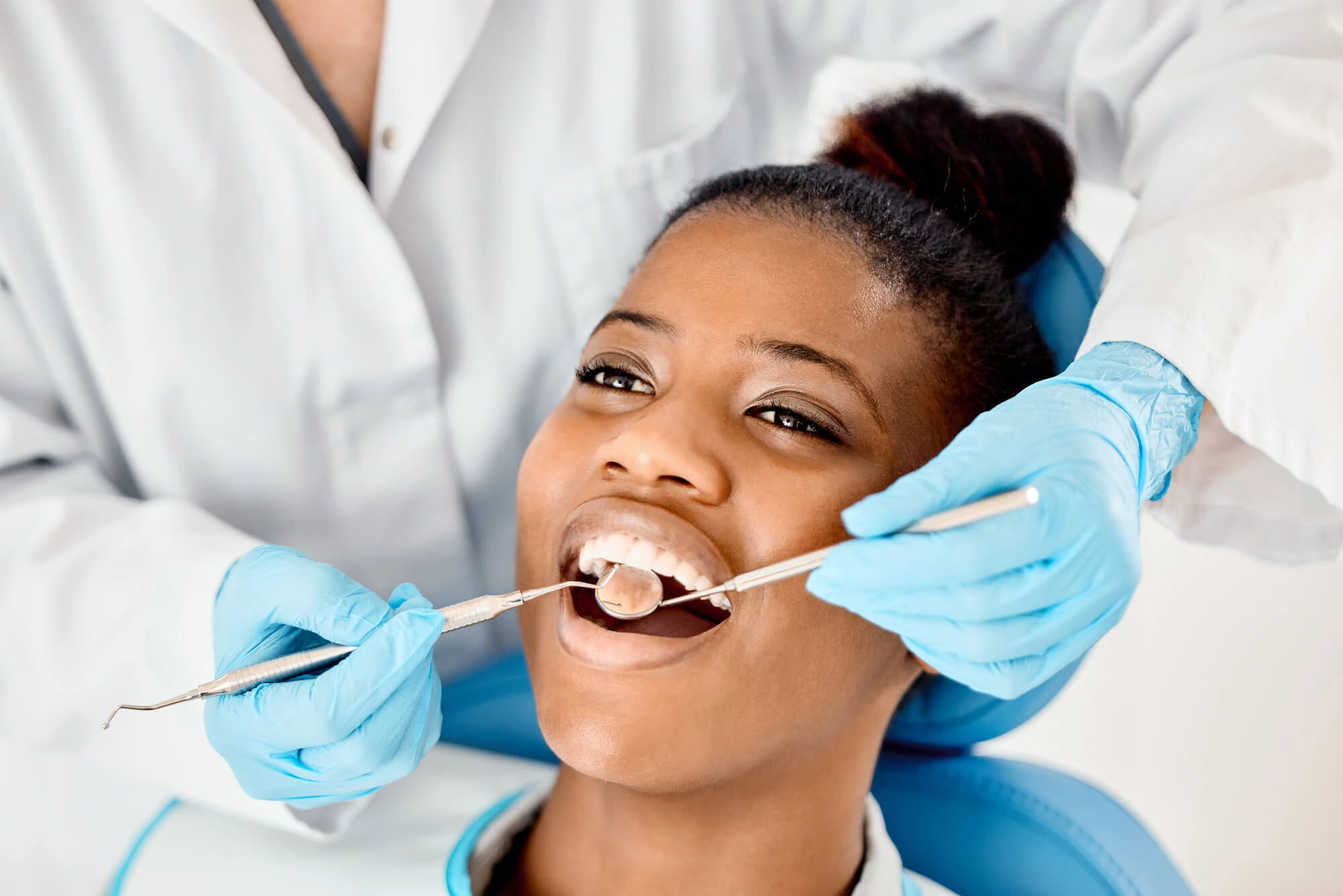 Dentist in white coat and blue gloves examining a smiling woman's teeth with dental tools.