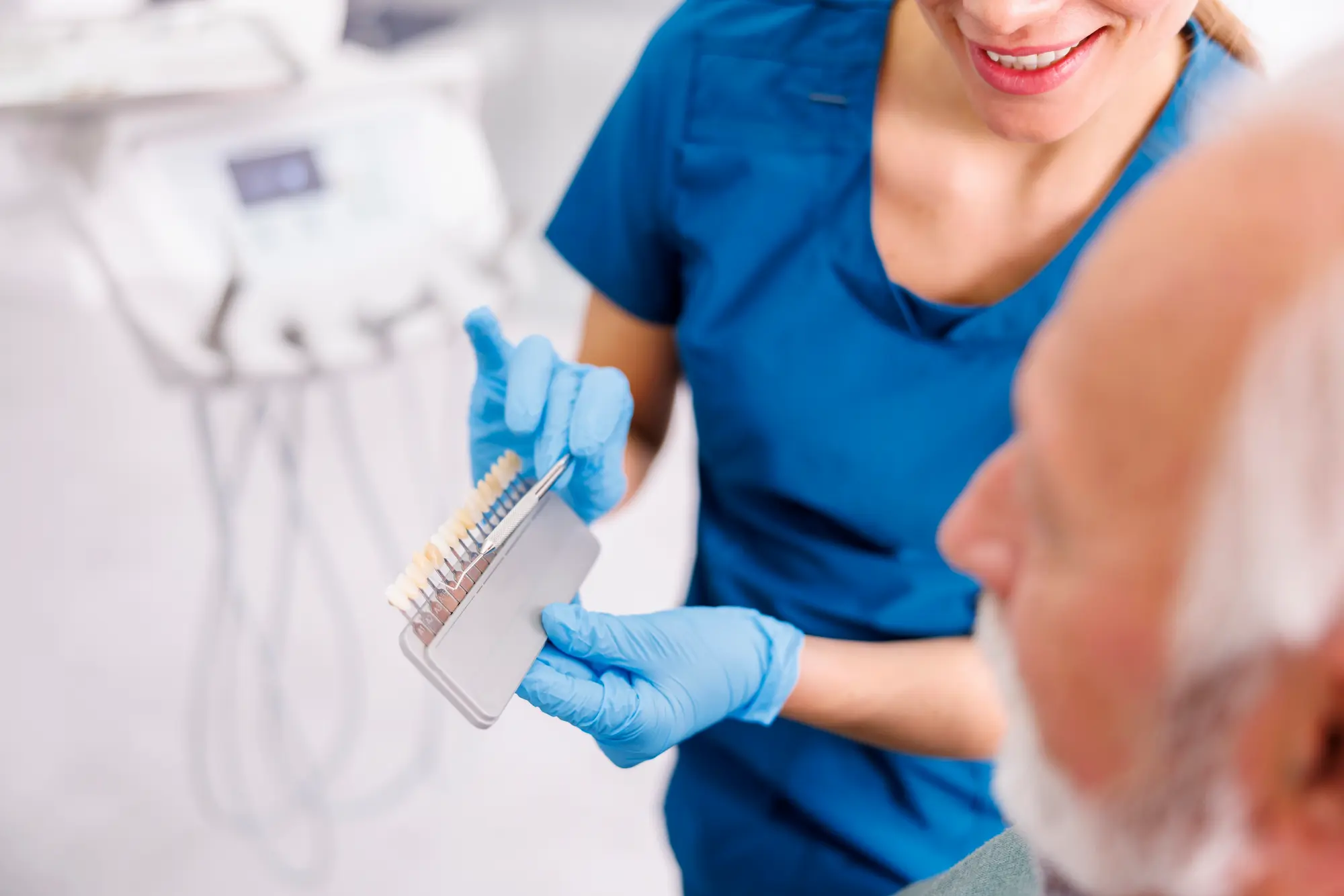 Dentist wearing blue scrubs and gloves showing a dental shade guide to an older male patient.