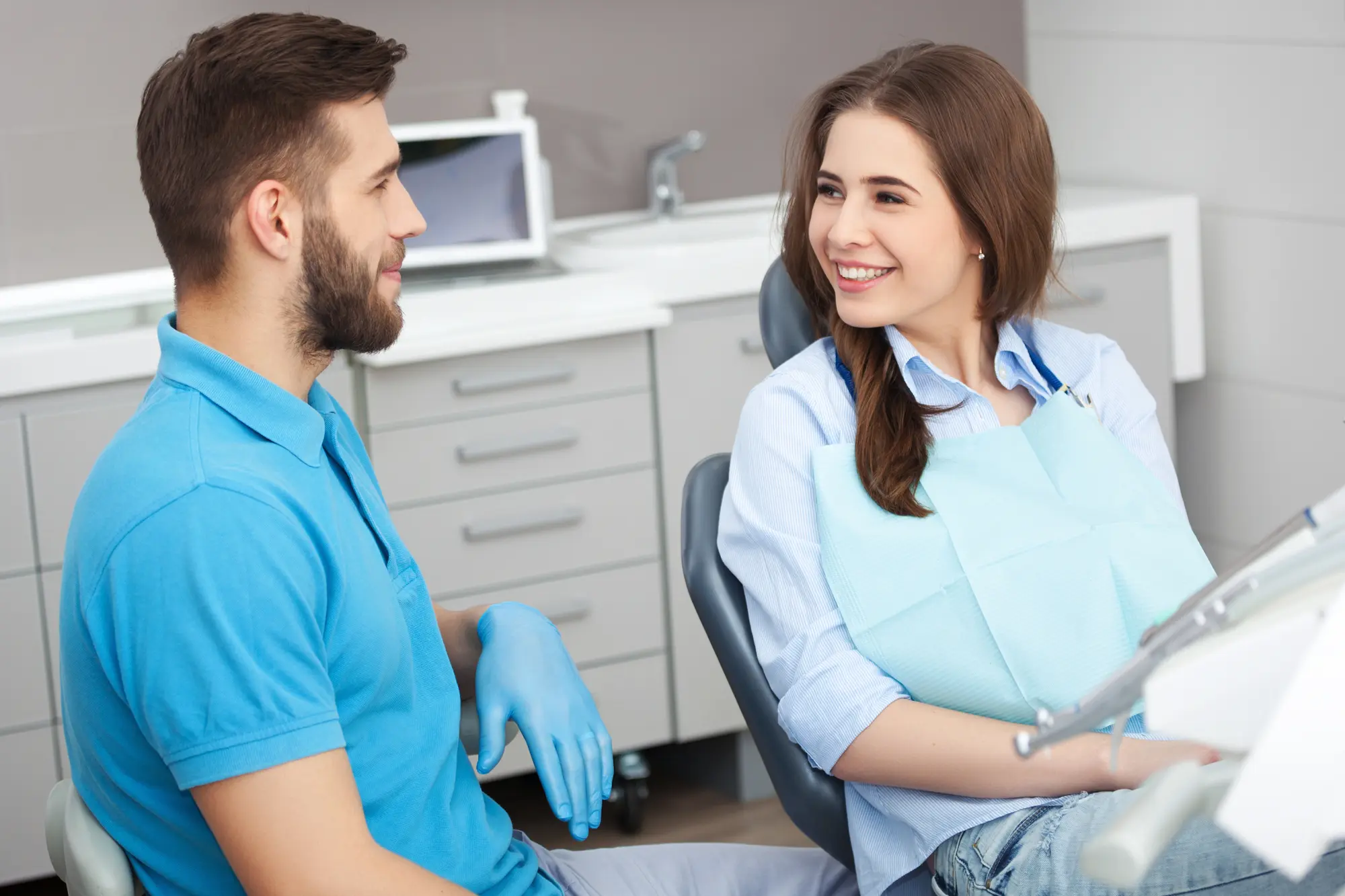 Smiling female patient sitting in a dental chair talking with a male dentist wearing blue gloves.