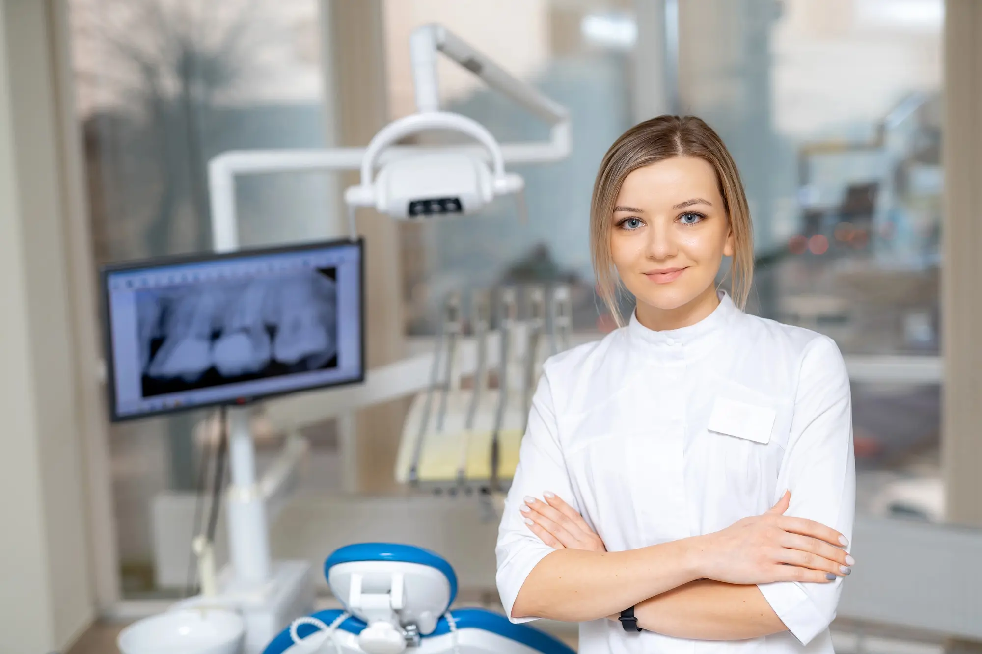 Female dentist standing with arms crossed in a dental clinic with an X-ray image of teeth displayed on a screen behind her.