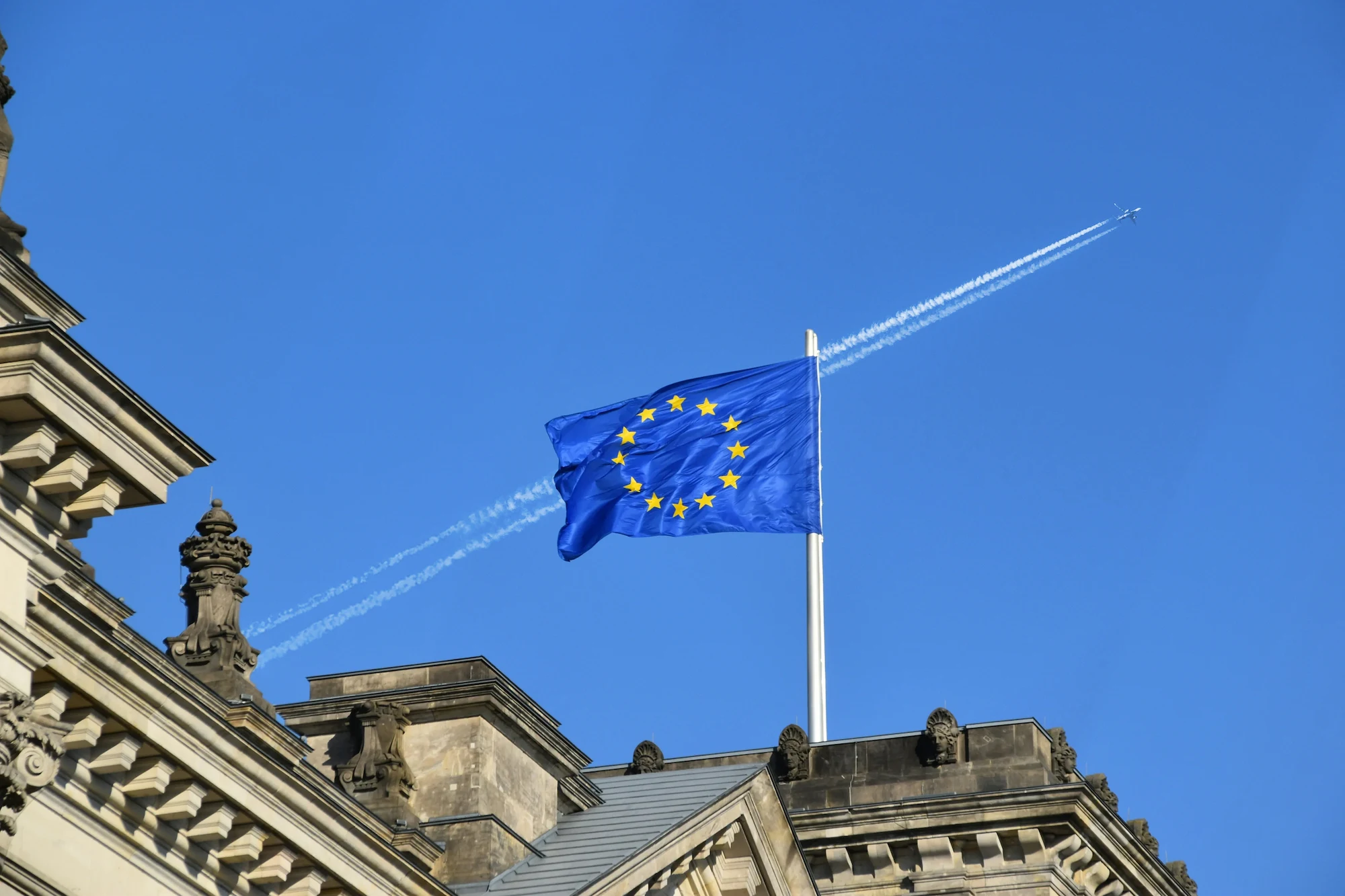 European Union flag waving atop a historic building with a plane and contrail in the clear blue sky.