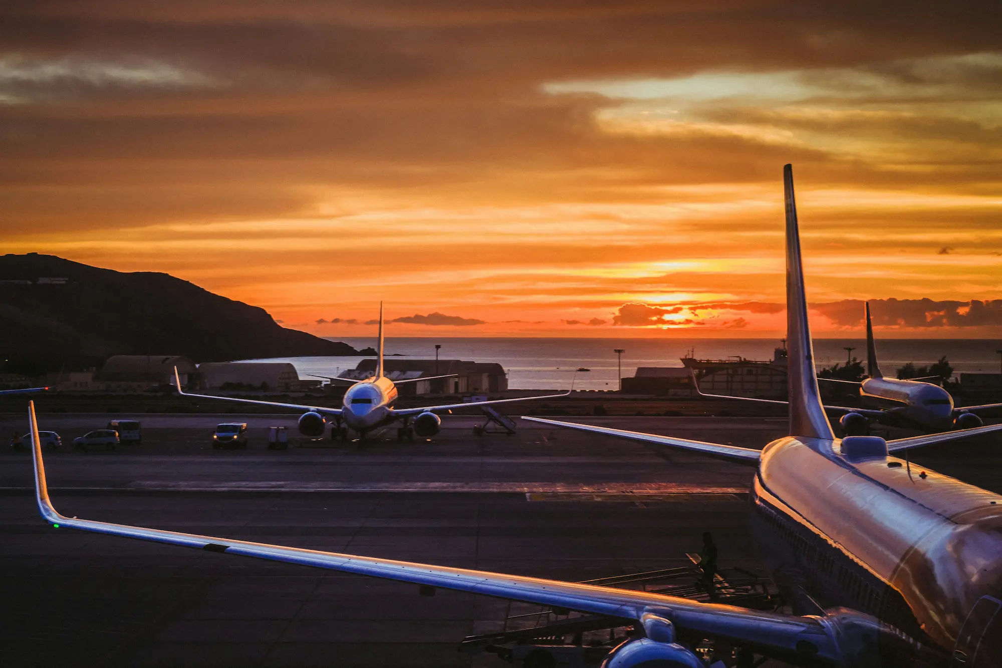 Airport runway with parked airplanes at sunset by the ocean and hills in the background.
