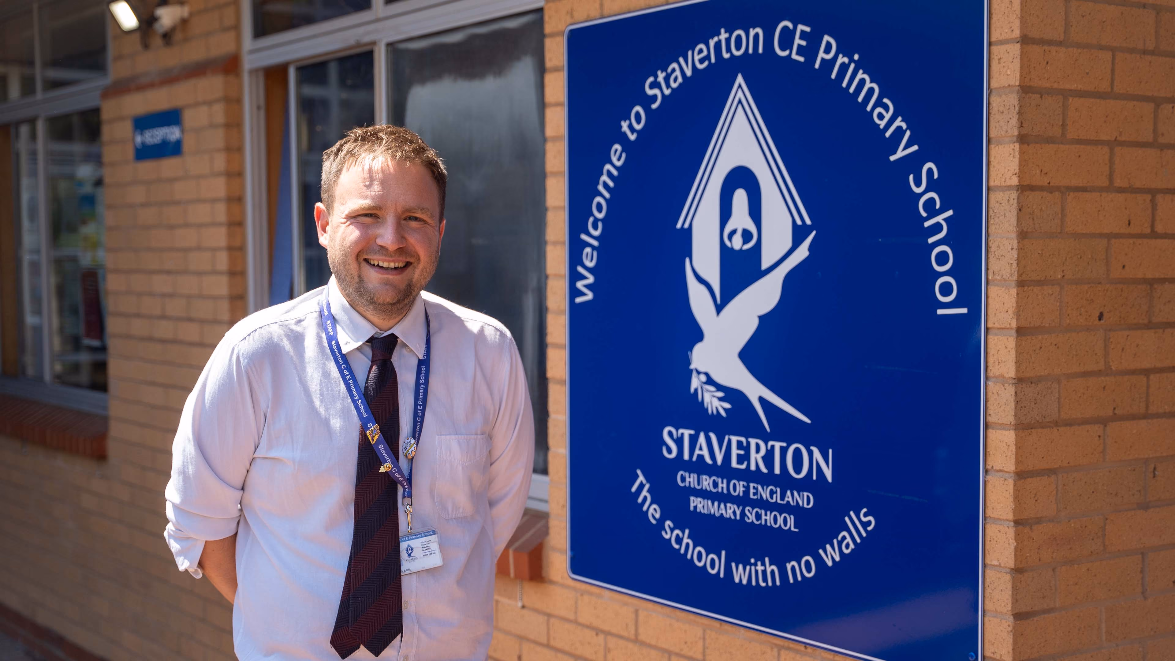 Smiling man in a white shirt and tie standing beside the Staverton CE Primary School sign.