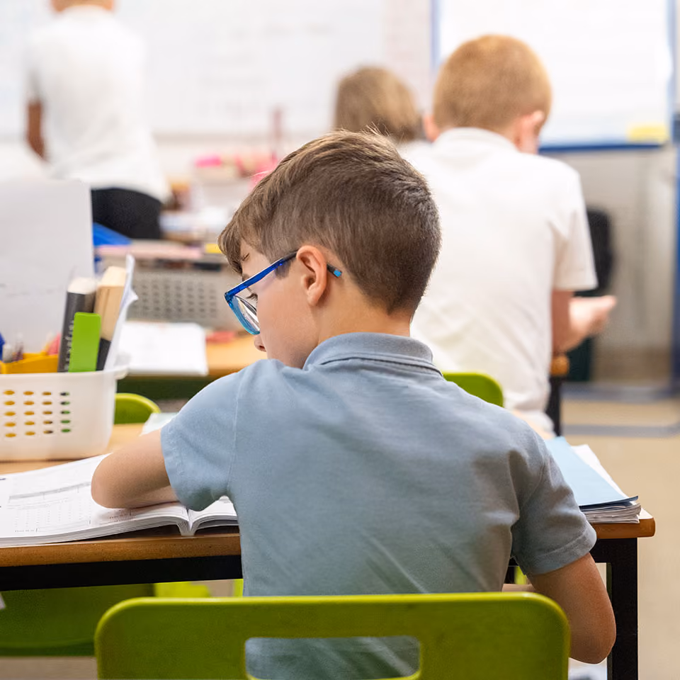 Young boy with glasses sitting at desk and writing in a workbook in a classroom.
