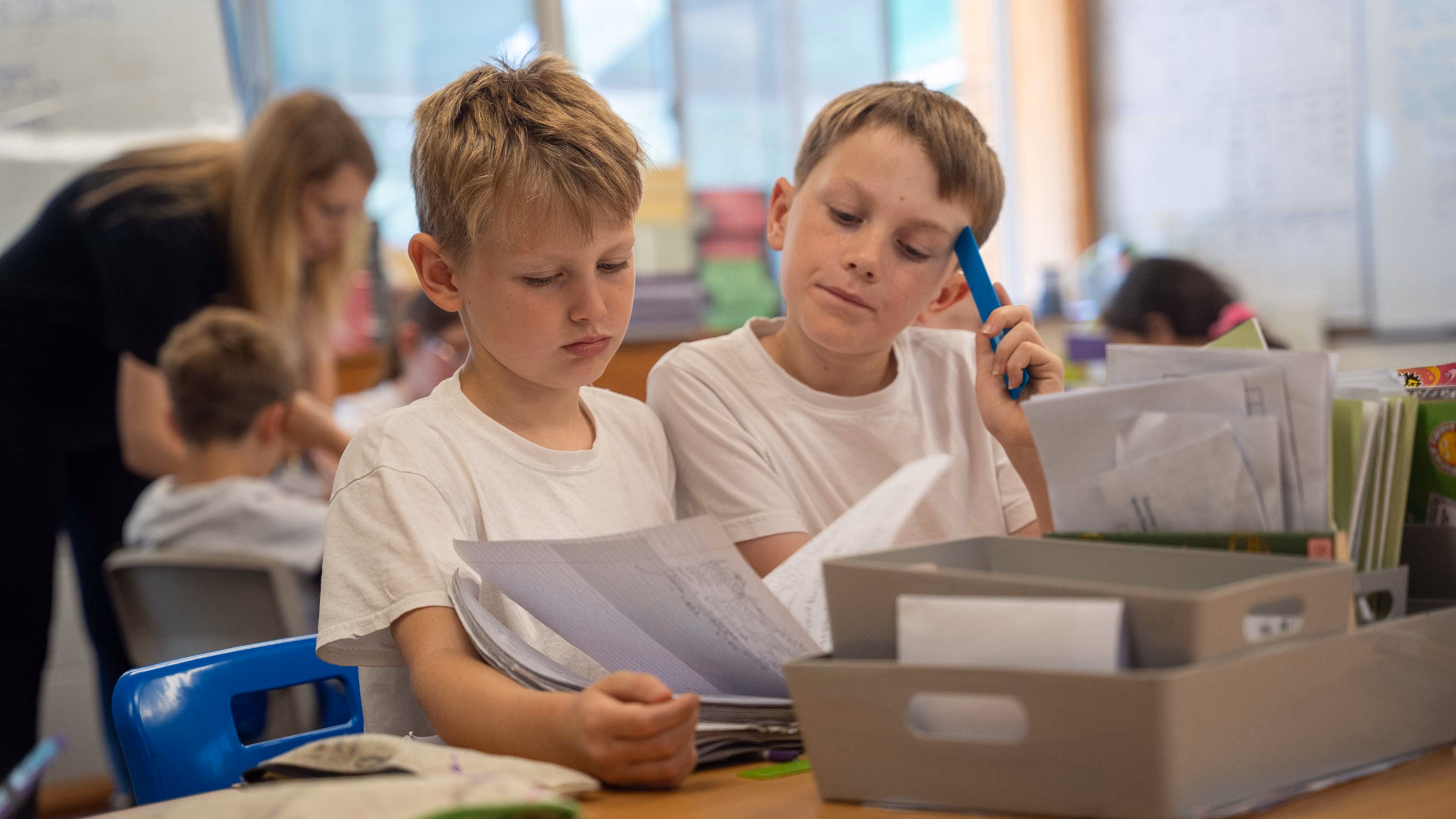 Two boys in white shirts reviewing papers together at a classroom desk with supplies.