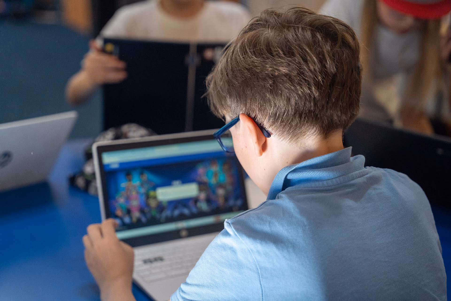 Child wearing blue shirt and glasses using a laptop at a table with others in background.