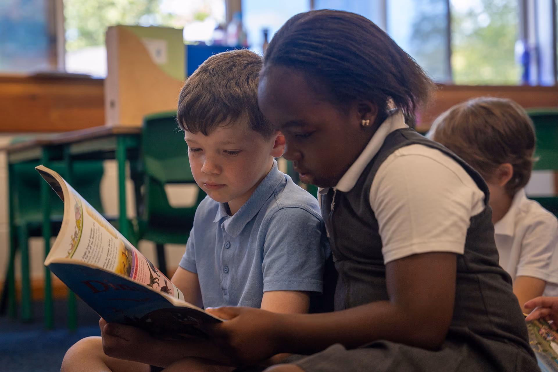 Two young children sitting closely and reading a book together in a classroom.