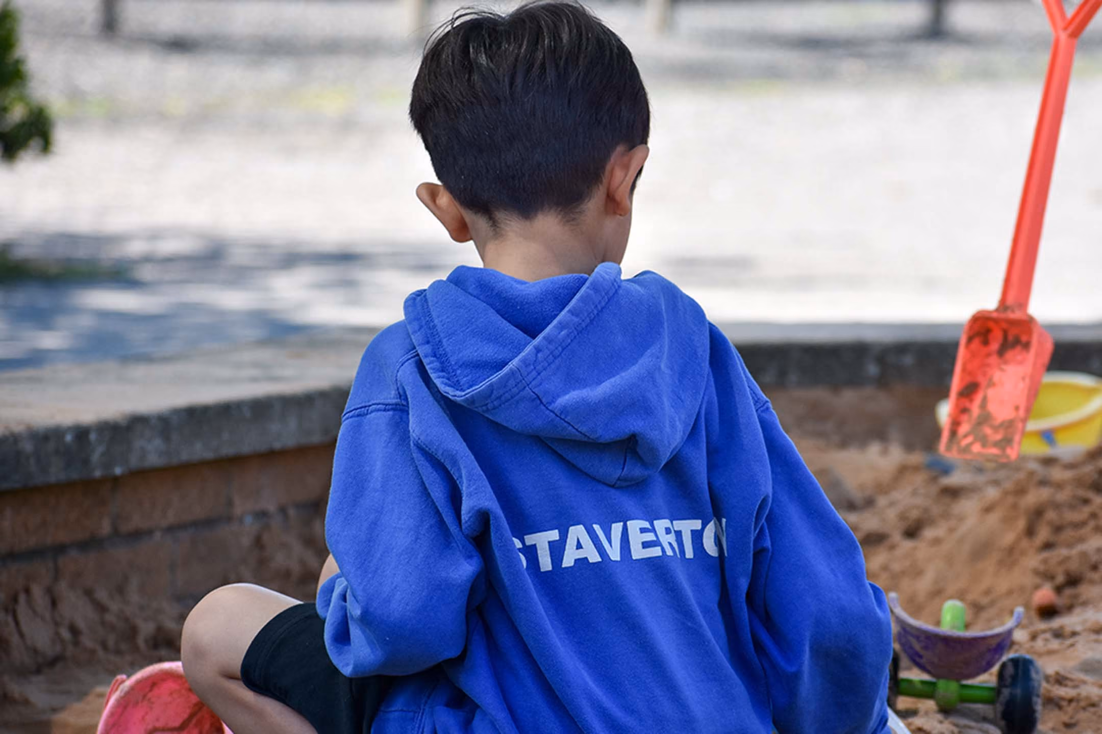 Child in a blue Staverton hoodie playing in a sandbox with toys, viewed from behind.