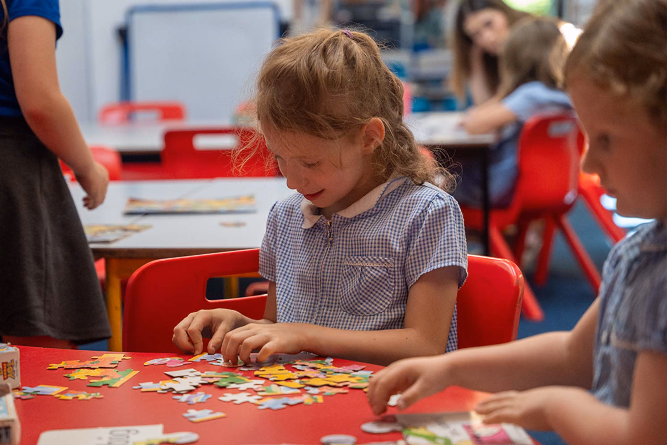 Young girl in a checkered dress assembling a colorful puzzle at a red table in a classroom.