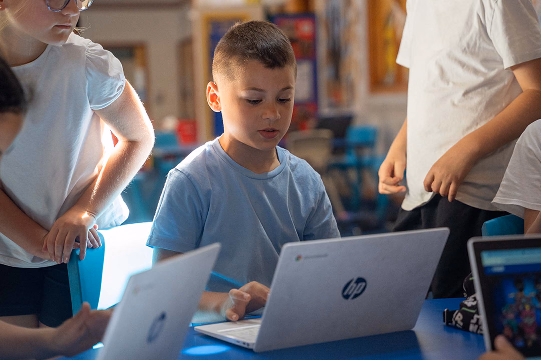 Young boy using an HP laptop at a table surrounded by other children in a classroom.