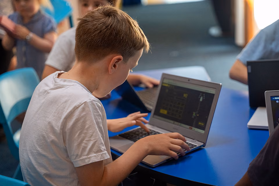 A boy in a white shirt using a laptop at a blue table with other children also on laptops.