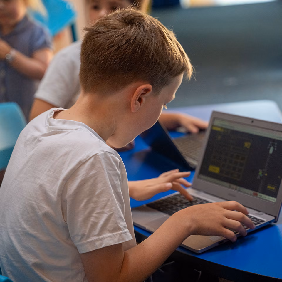 A boy in a white shirt using a laptop at a blue table with other children also on laptops.