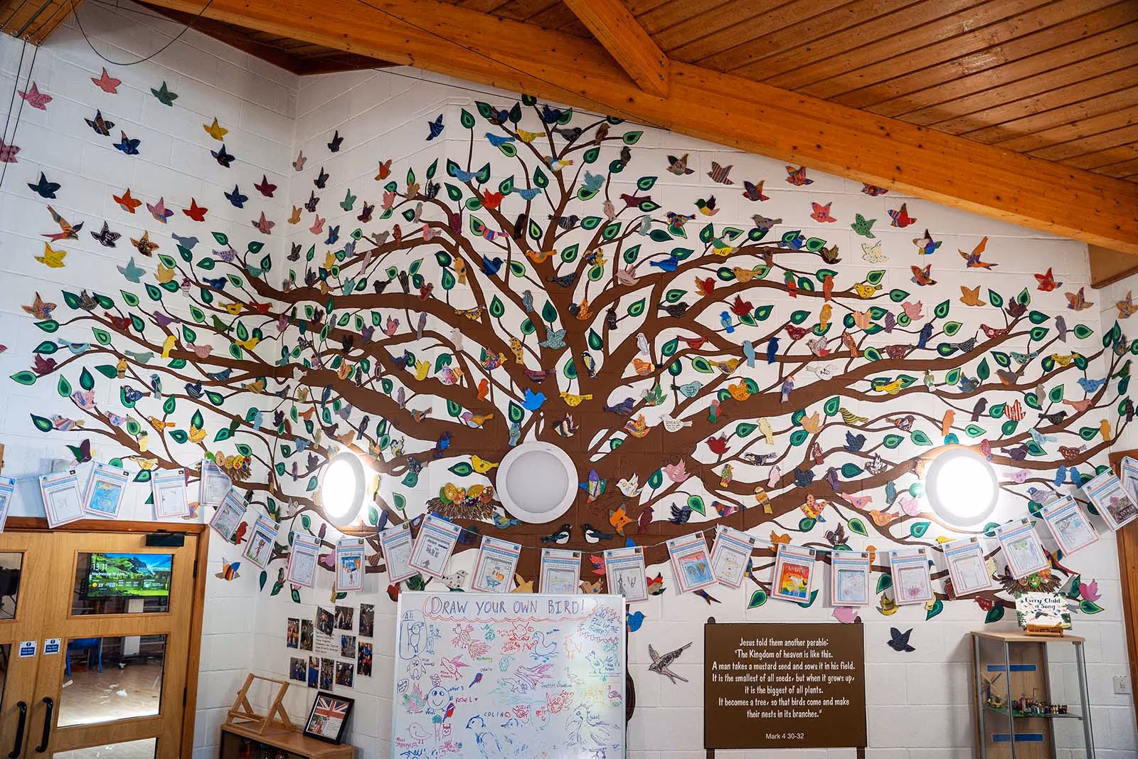 Wall mural of a tree with colourful paper birds and leaves, a whiteboard, and a Bible quote.
