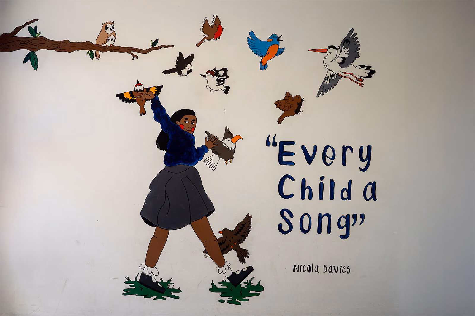 Wall mural of a girl surrounded by colourful birds, with the quote 'Every Child a Song' by Nicola Davies.