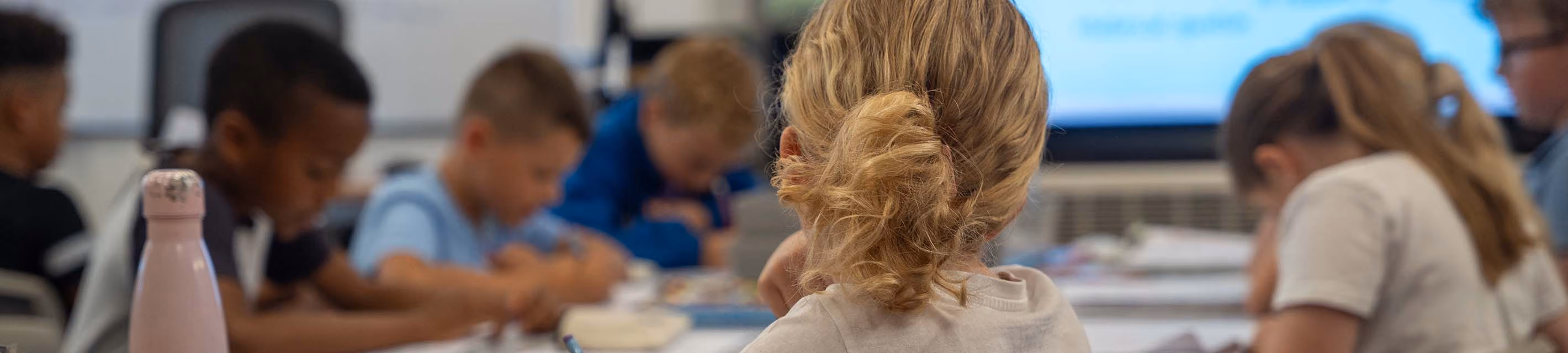 Pupils seated at desks working in a classroom, viewed from behind.