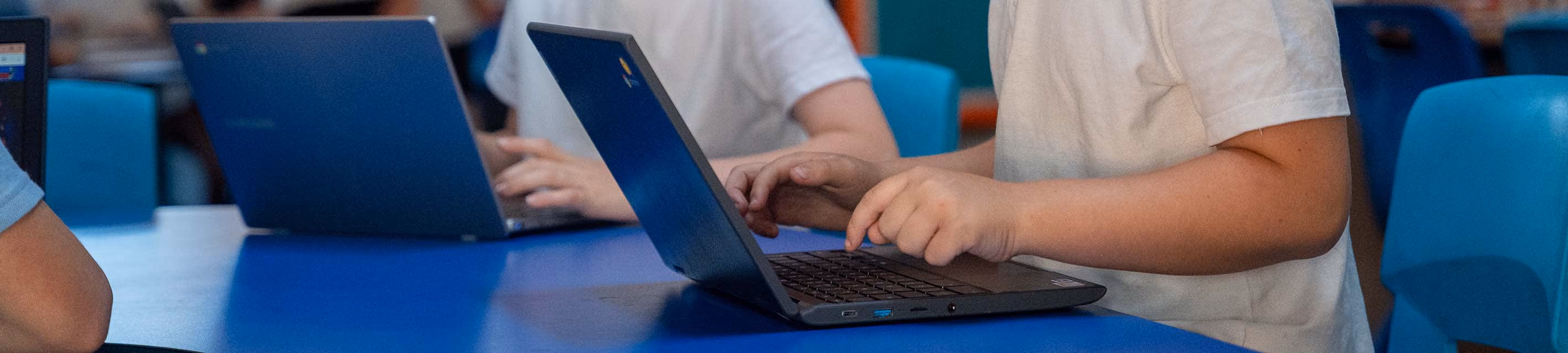 Two sets of boys hands typing on their laptops while sitting at a blue table in a classroom.