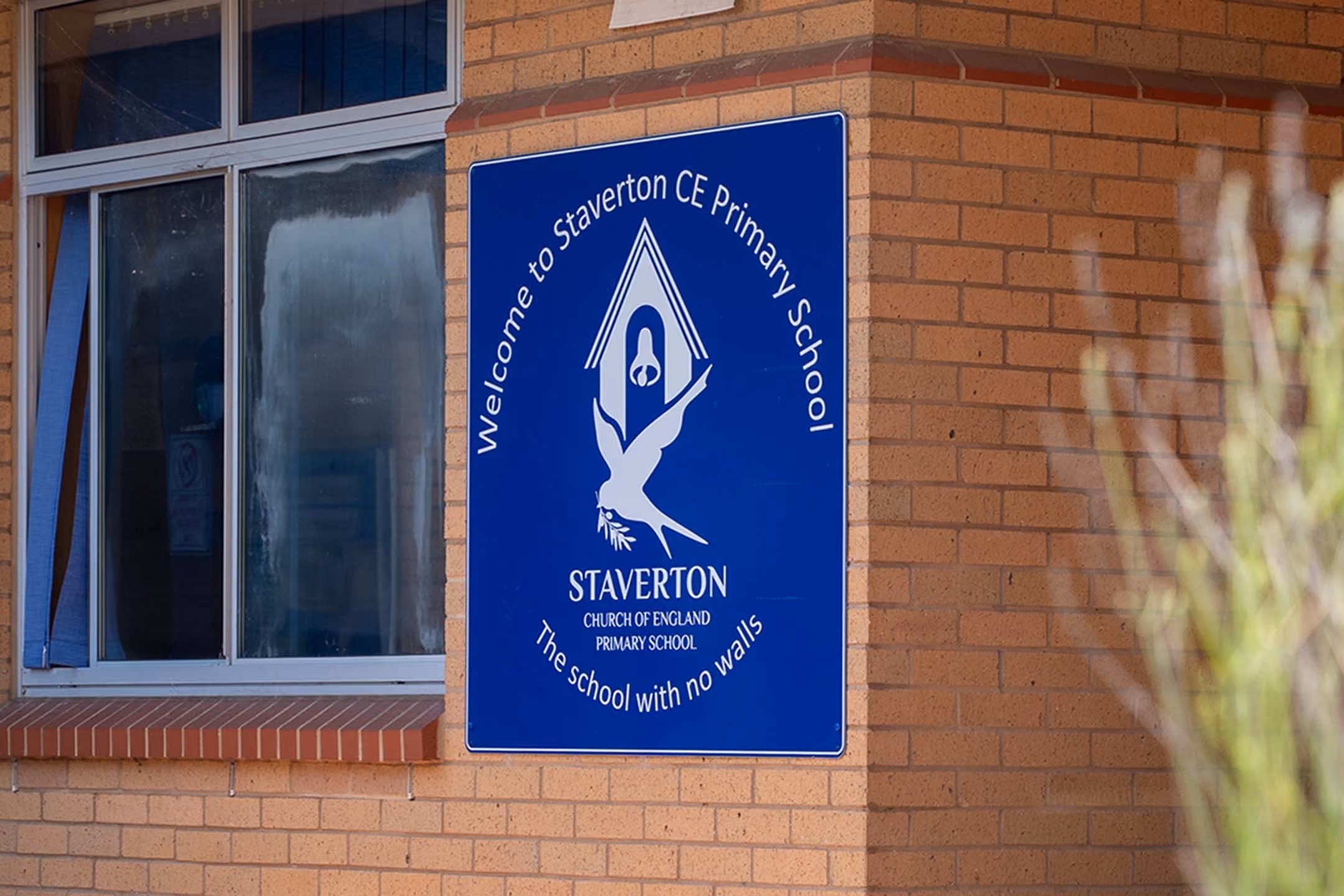 Blue welcome sign for Staverton Church of England Primary School on a brick building.