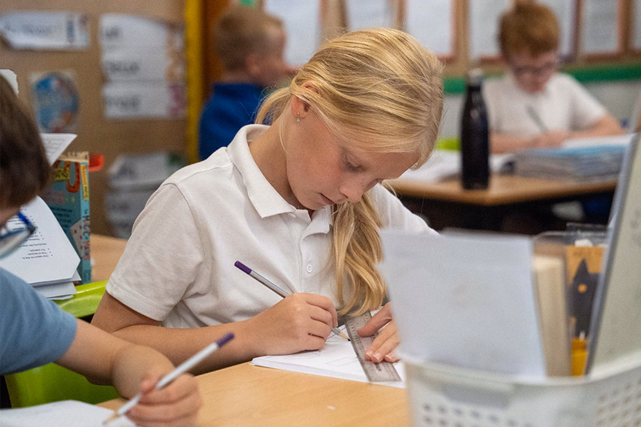 Pupil in school uniform using a ruler and pencil to write in an exercise book at her desk.