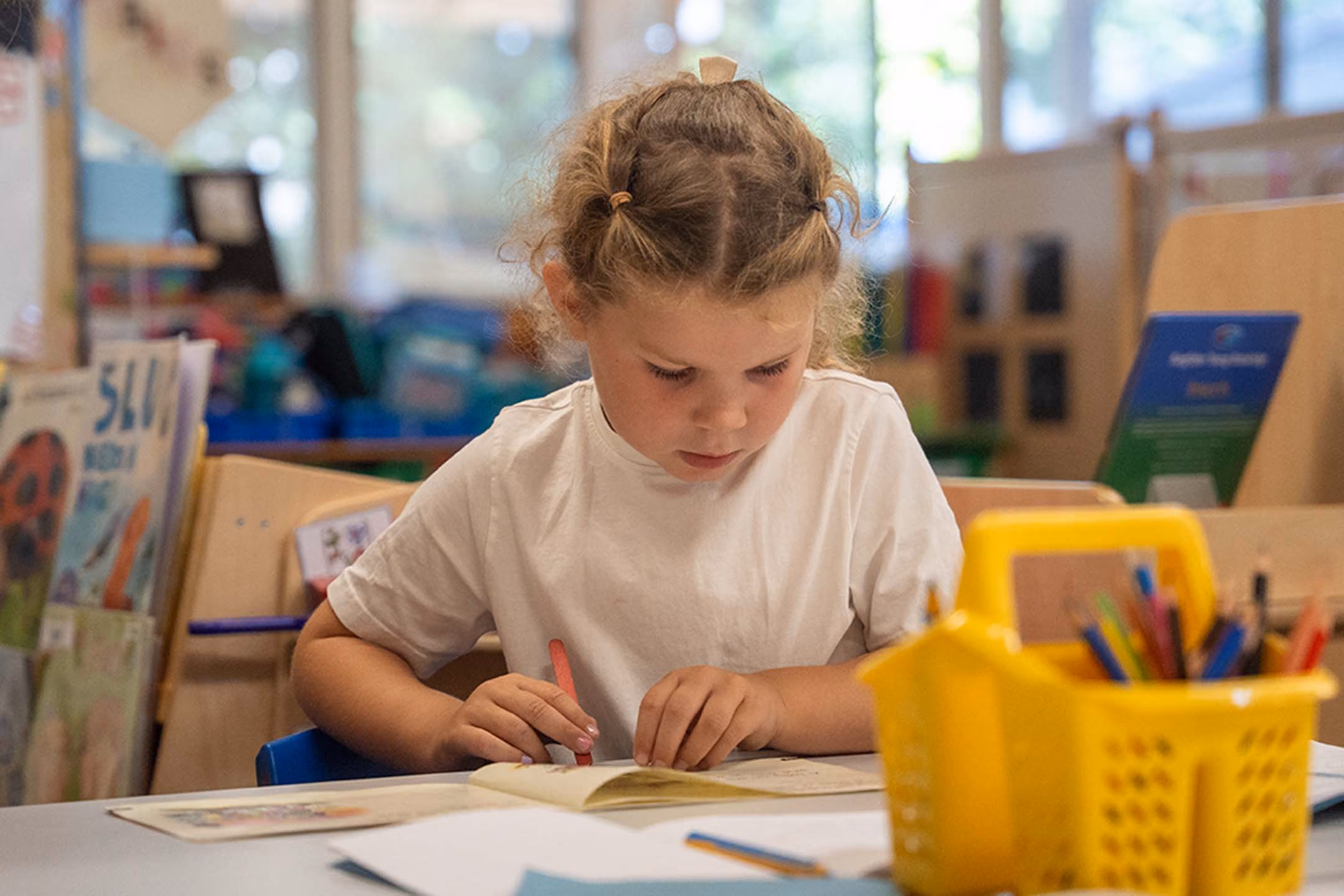 Young pupil writing in a book at her classroom table, with a pot of coloured pencils nearby.