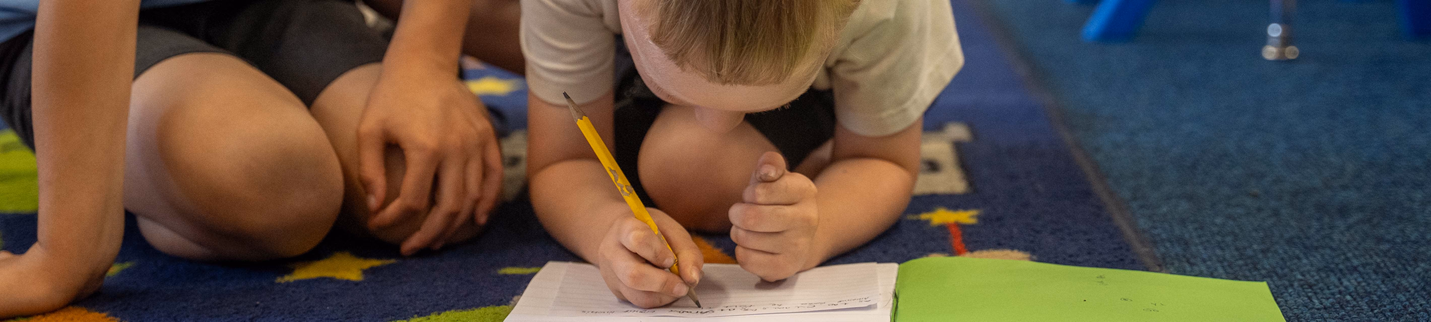 Two young boys on a classroom rug with one writing in a notebook and the other watching.