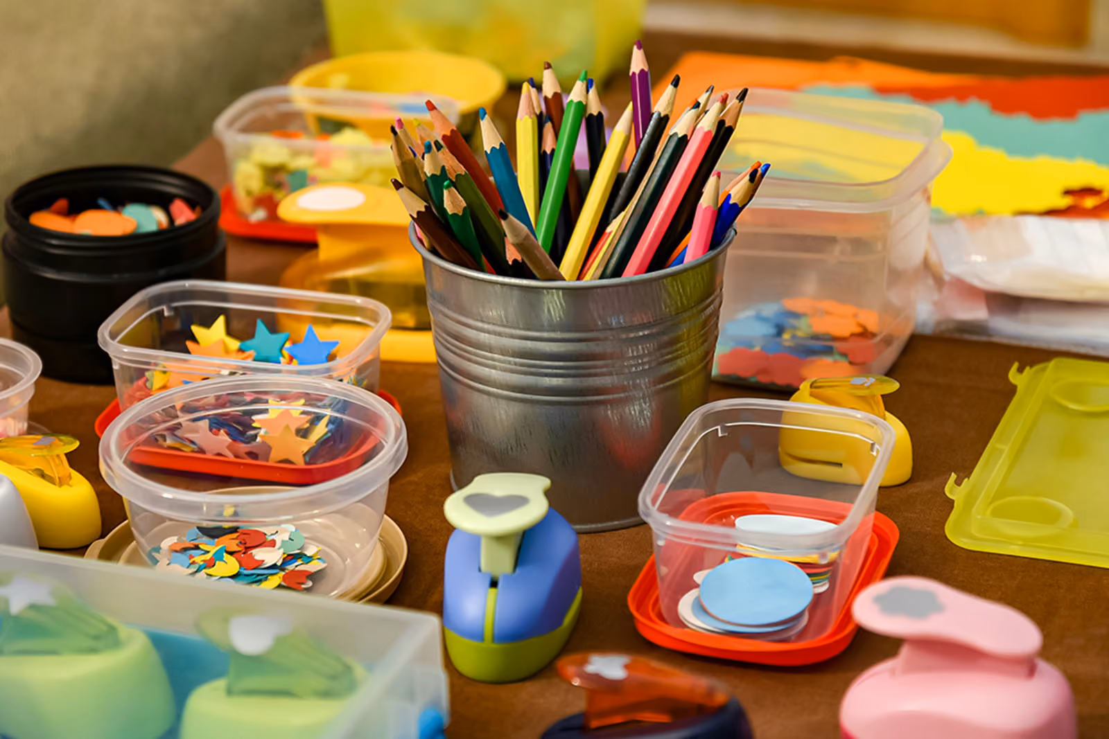 Colouring pencils in a metal pot surrounded by craft supplies, including shape punches and containers of paper shapes.
