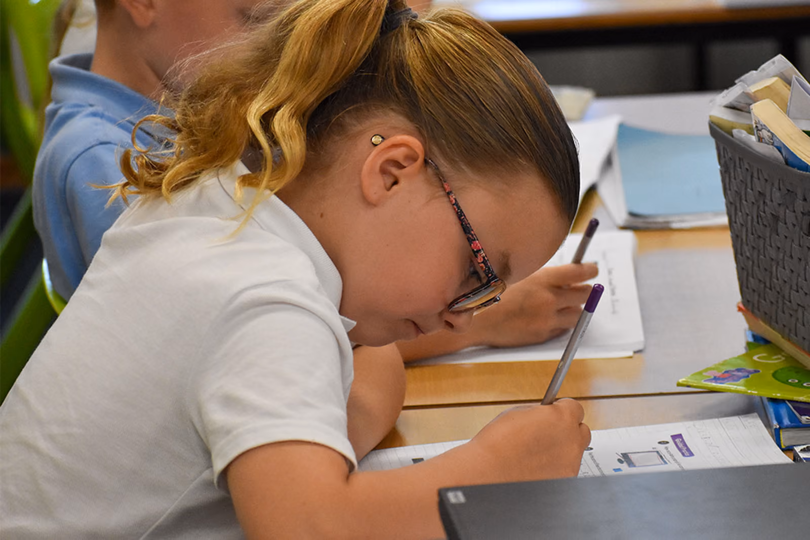 Pupil in glasses bent low over her desk, writing closely on paper.