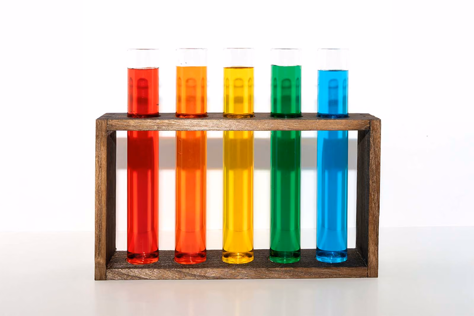 Five test tubes filled with red, orange, yellow, green and blue liquid in a wooden rack.